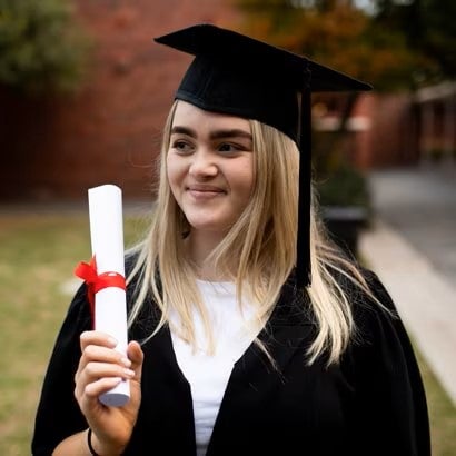 A smiling woman holds her degree while dressed in a graduation cap and gown