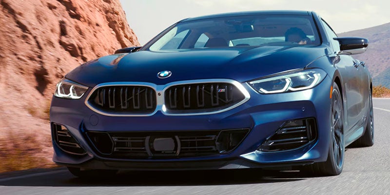 Blue BMW coupe driving on a road beside rocky canyon cliffs, shown from a front-angle perspective highlighting the vehicle's grille, headlights